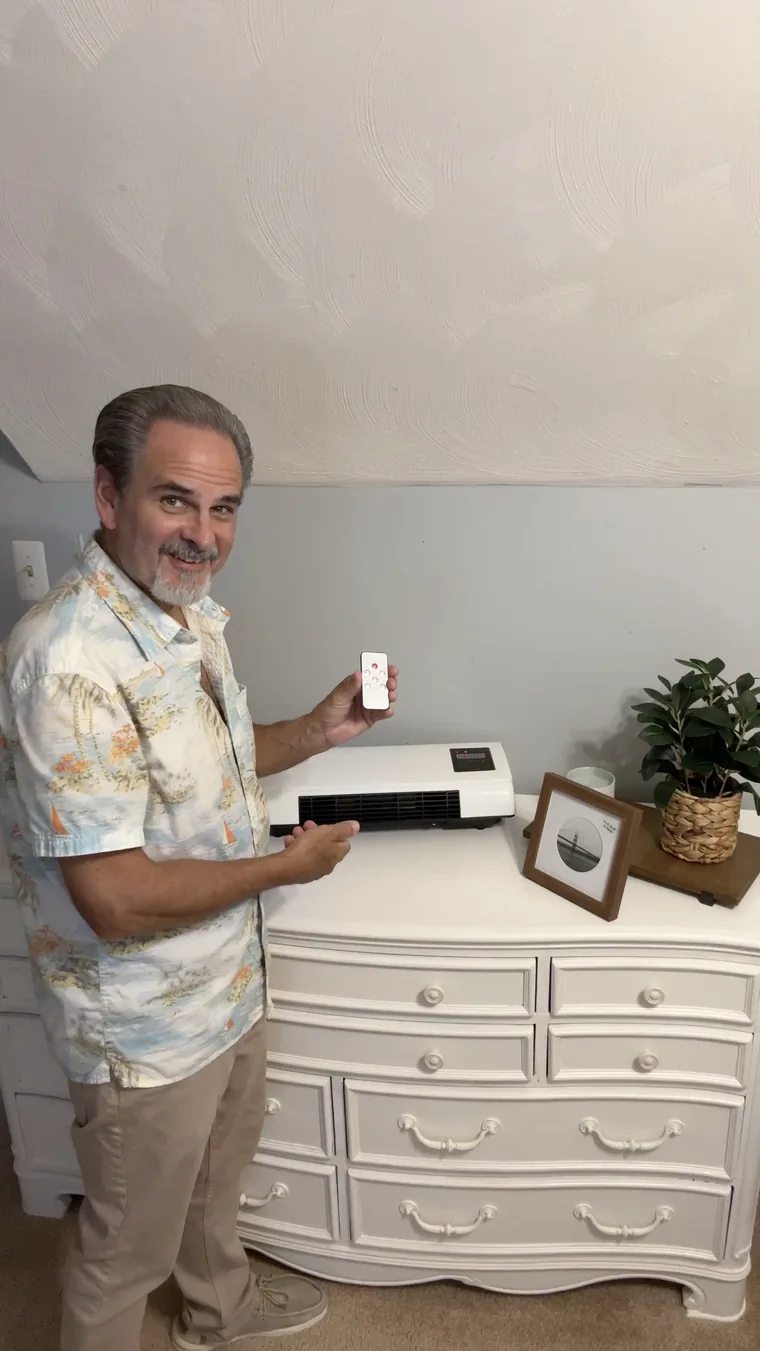 A man holding the small remote next to the heater on a dresser